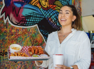 woman eating hot chicken sandwich and fries in front of mural 